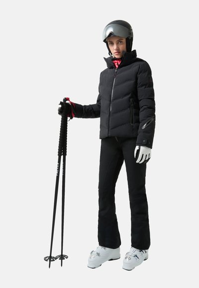 Schwarze Pufferjacke, enganliegende schwarze Skihose, weiße Skischuhe mit silbernen Akzenten, hält schwarze Skistöcke, trägt einen grauen Helm und Handschuhe.