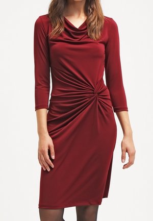Kobieta w bordowej sukience do kolan z długimi rękawami, z marszczonym detalem zawiązanym w talii i dekoltem z fałdami.