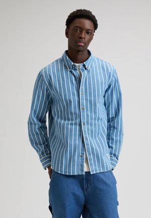 Młody mężczyzna ubrany w niebieską koszulę zapinaną na guziki z białymi pionowymi paskami na białym T-shircie i nieformalnych niebieskich dżinsach, stojący na jednolitym tle.