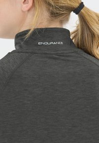 Blonde Person mit Pferdeschwanz, trägt ein dunkelgraues Sporthemd mit hohem Kragen, auf dem Rückenkraagen steht der Text "ENDURANCE".