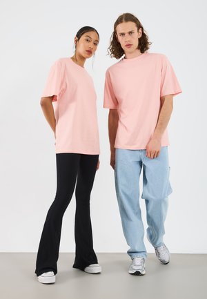 To modeller iført matchende pastellrosa t-skjorter. Den ene kombinerer det med svarte ribbede flaredbukser, mens den andre har på seg lyseblå baggy jeans.