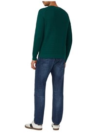 Groen gebreide pullover met ronde hals, gedragen over blauwe spijkerbroek en witte sneakers met zwarte accenten. Textuurdetails zichtbaar op de stof.