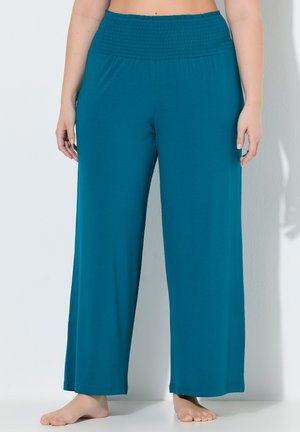 Vrouw die een losse teal broek met hoge taille draagt en blootsvoets staat tegen een witte muur, waarbij de pasvorm en details van de tailleband van de broek zichtbaar zijn.