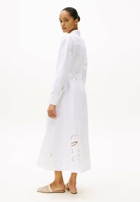 Vestido blanco de manga larga hecho de tela ligera, con cintura ajustada y bordado floral en el dobladillo. Combinado con zapatillas discretas de color neutro.