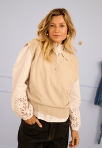 Bézs kötött pulóvermellény gallérral és elöl gombos részlettel, fehér blúz felett, amely hímzett mandzsetta díszítéssel rendelkezik. Sötét farmernadrág.