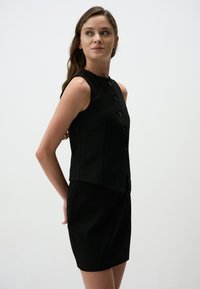 Robe noire sans manches avec un corsage ajusté, un col montant et cinq boutons noirs sur le devant ; texture lisse et jupe courte.