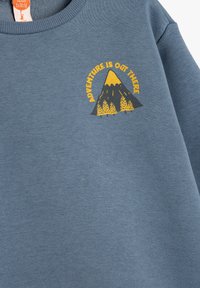 Niebieska bluza z okrągłym dekoltem, z grafiką góry i napisem "PRZYGODA CZEKA NA CIEBIE" w żółtym i czarnym kolorze. Miękka tekstura.