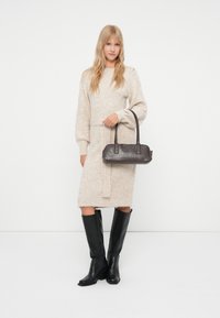 Abito maglione beige con maniche lunghe e vita legata, abbinato a stivali neri in pelle fino al ginocchio e a una piccola borsa scura.