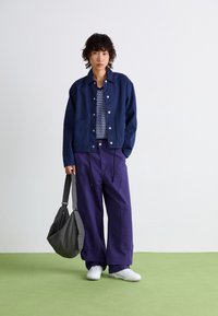 Veste bleue marine à boutons avec deux poches avant, associée à un débardeur à pois et à un pantalon large violet. Tenant un sac à carreaux noir et blanc.