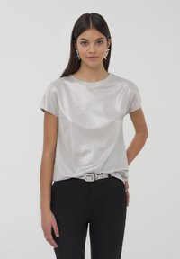 T-shirt argento a maniche corte con una texture lucida e riflettente. Vestibilità ampia, scollatura rotonda, abbinata a pantaloni neri e una cintura con strass.