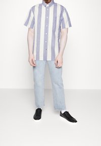 Koszula z krótkim rękawem z pionowymi paskami w kolorze lawendy i białym, kołnierzyk, guziki, pojedyncza kieszeń na piersi; połączona z jasnoniebieskimi jeansami i czarnymi trampkami.