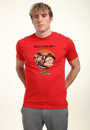 Ung mand iført en rød t-shirt med Street Fighter-grafik, der viser to figurer i kampstilling og japansk tekst ovenover.