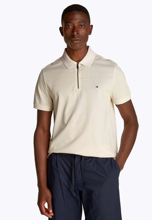 Cremefarbenes Poloshirt mit Reißverschlusskragen, das ein strukturiertes Streifendesign am Kragen und ein kleines Logo auf der Brust aufweist. Kombiniert mit dunkler Hose.