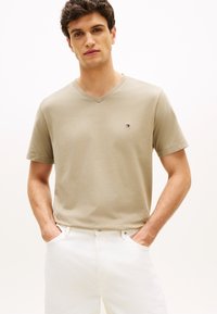 Μπεζ T-shirt με λαιμόκοψη V, κατασκευασμένο από μαλακό ύφασμα με κοντά μανίκια, που διαθέτει μικρό, κεντητό λογότυπο στο στήθος, σε συνδυασμό με λευκό παντελόνι.