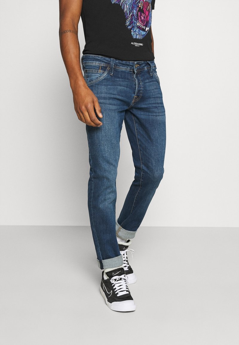 Man med blå upprullade jeans, svart grafiskt t-shirt och svarta och vita Nike-sneakers som står mot en enkel ljus bakgrund.