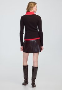 Kobieta odwrócona plecami, ubrana w czarną bluzkę z długim rękawem i czerwonym kołnierzykiem, ciemno-brązową skórzaną spódnicę oraz ciemno-brązowe kozaki za kolano.