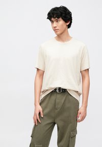 Béžové tričko s kulatým výstřihem z lehké tkaniny, doplněné olivově zelenými kalhotami s bočními kapsami a černým pásem s průchodkami.