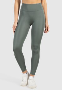 Zelené legíny na cvičení s vysokým pasem vyrobené z pružné, hladké tkaniny. Přiléhavý střih, bezešvá konstrukce, doplněné o bílé sportovní tenisky.
