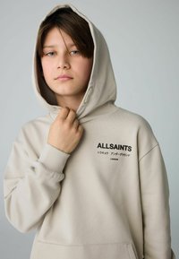 Sudadera color beige con un bolsillo frontal y capucha ajustable. Presenta el logo "ALLSAINTS" en negro, fabricada con un tejido liso y texturizado.