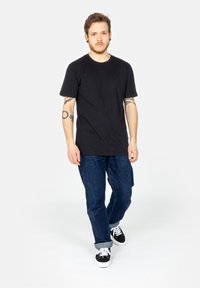 Schwarzes Baumwoll-T-Shirt mit kurzen Ärmeln, kombiniert mit dunkelblauen Jeans und schwarzen Sneakers, stehend vor einem schlichten weißen Hintergrund.