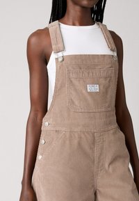 Beige corduroy overalls with adjustable shoulder straps, metal hardware, front pocket, and side button accents. Worn over a white top.