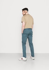 Chemise beige à manches courtes, pantalons ajustés teal, et baskets blanches avec des accents rouges. La personne est vue de dos contre un fond blanc.