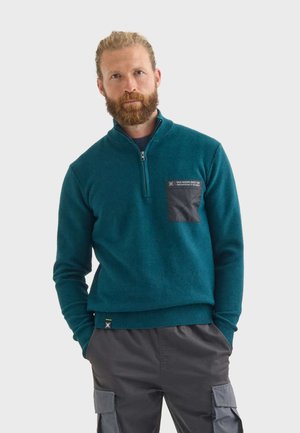 Pullover en polaire teal avec un col zippé, featuring une poche poitrine noire et un tissu texturé. La coupe est droite avec des poignets et un ourlet côtelés.