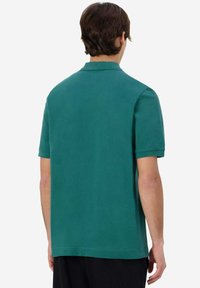 Polo shirt turchese a maniche corte realizzato in cotone. Presenta un colletto classico e una vestibilità rilassata con una texture semplice e uniforme. Nessun motivo visibile.