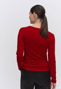 Långärmad topp i röd sammet med rund halsringning och figurnära design, med en rynkad textur längs ryggen. Svarta byxor är på.
