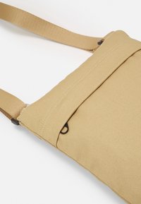 Strikstof crossbody tas i beige, med en frontlomme med en sort lynlås, justerbar rem og flad rektangulær form.