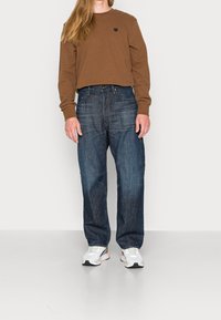 Brun långärmad tröja, mörkblå denimjeans och vita sportskor. Avslappnad passform med en liten märkeslapp på tröjan.