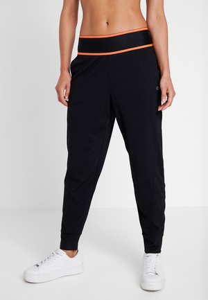 Femme portant un pantalon jogging noir avec une taille bordée d'orange et des baskets blanches, debout devant un fond uni clair.