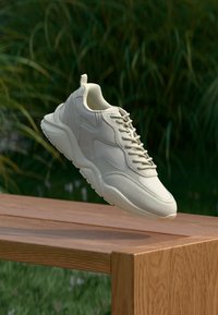 Béžová teniska so šnúrkami vznášajúca sa nad drevenou lavičkou vonku, v pozadí rozmazané zelené rastliny.