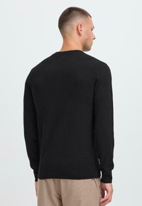 Man die zijwaarts kijkt in een effen zwart langemouwen trui en lichtbeige broek voor een effen lichte achtergrond.
