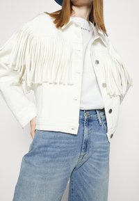 Chaqueta de mezclilla blanca con detalles de flecos, cierre de botones y dos bolsillos frontales. Lleva puesta sobre una camisa blanca y combinada con jeans de color azul claro.