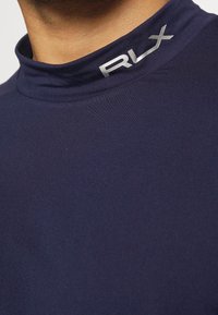 Detailní záběr na límec námořnického modrého roláku s stříbrným logem "RLX" na levé straně.