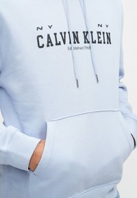 Svetlo modrá mikina s kapucňou, so prednou kapsou a šnúrkami. Zobrazuje text "CALVIN KLEIN" a "Established 1968" v čiernej farbe.