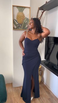 Ženska v dolgi temno modri obleki s perlastimi naramnicami stoji nasmejana, z roko za glavo, blizu televizorja in okvirja s sliko ananasa na steni.