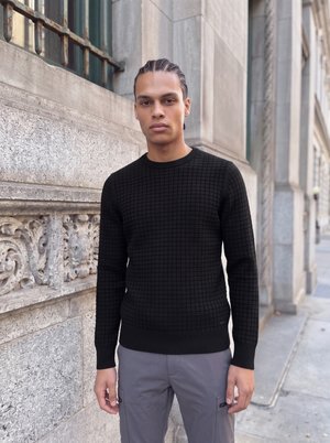 Un homme aux cheveux tressés se tient sur un trottoir en ville, à côté d’un bâtiment en pierre sculptée. Il porte un pull noir à la texture travaillée et un pantalon gris.