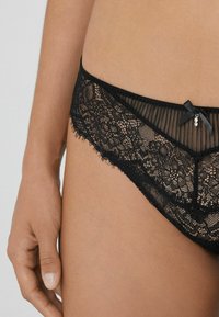Lingerie en dentelle noire avec des détails en tulle transparent, présentant un motif floral, des bords festonnés et un petit nœud noir avec un accent en gemme.