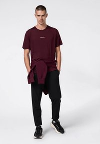 Burgunderrotes T-Shirt mit kurzen Ärmeln und einem Logo auf der Brust, kombiniert mit schwarzen Jogginghosen und einem Sweatshirt zum Umschlagen. Verfügt über eine lockere Passform.