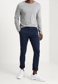 Šedé tričko s dlouhým rukávem, modré chino kalhoty vyhrnuté na manžetách a bílé tenisky. Jednoduchý design s volným střihem a hladkými texturami.