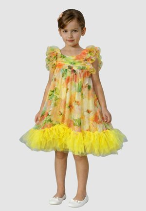 Ragazza giovane in piedi, che indossa un vestito floreale colorato con maniche arricciate e un orlo di tulle giallo brillante, e ballerine bianche.