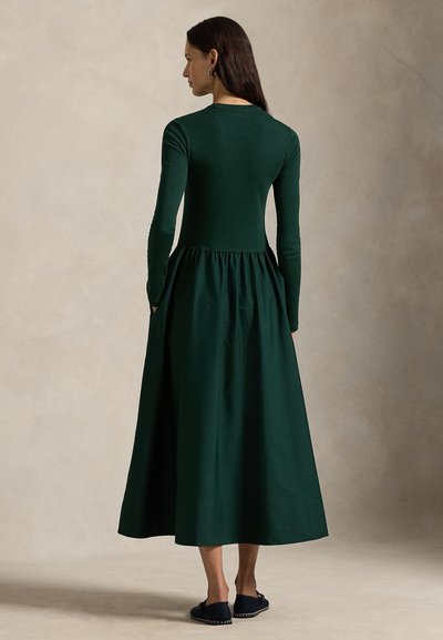 Mörkgrön bomulls-klänning med lång ärm, figursydd överdel och en rynkad kjol som når till mitten av vaden. Bärs med svarta slip-on skor.