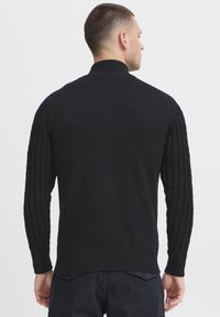 Zwarte gebreide trui met een hoge kraag en kabelpatroon op de mouwen. Heeft een gladde torso en een geribbelde zoom.