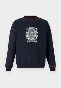 Sudadera azul marino con costuras rojas. Presenta un gran emblema blanco con el texto "Hilfiger" y elementos decorativos. Material de algodón y cuello redondo.