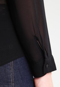 Chemisier noir transparent avec un poignet boutonné et un détail fendu. Il a une texture légère et est associé à un jean foncé.