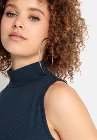 Marineblaue, gerippte ärmellose Bluse mit hohem Kragen. Das Model trägt große silberne Creolen. Die Haut hat einen natürlichen Glow und weiche Locken.