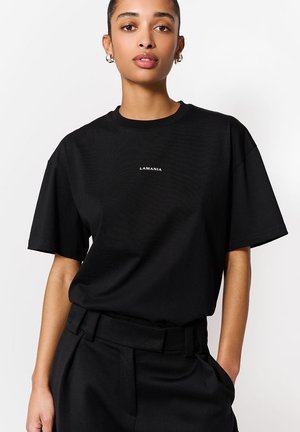 Kobieta o ciemnych włosach, nosząca srebrne kolczyki koła, prezentuje czarną koszulkę z krótkim rękawem z napisem "LAMANIA" oraz czarne spodnie z wysokim stanem.