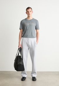 Pelēks T-kreklis ar "HUGO" logotipu, apvienots ar gaiši pelēkiem sporta biksēm. Melna soma vienā rokā, melnas kurpes. Minimalistisks dizains ar mīkstām tekstūrām.